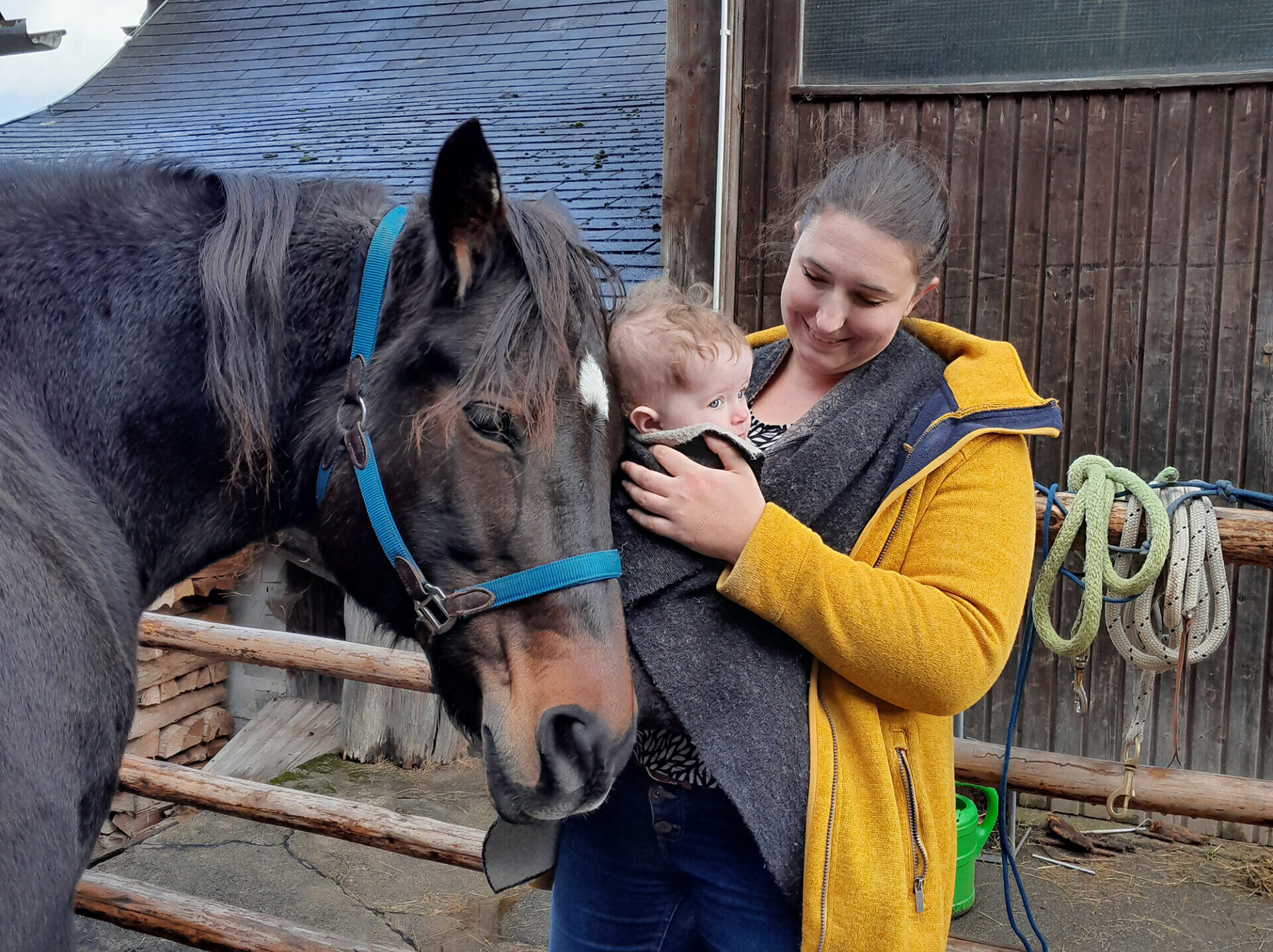 Mutter mit Baby im Tragetuch neben einem Pferd am Stall.